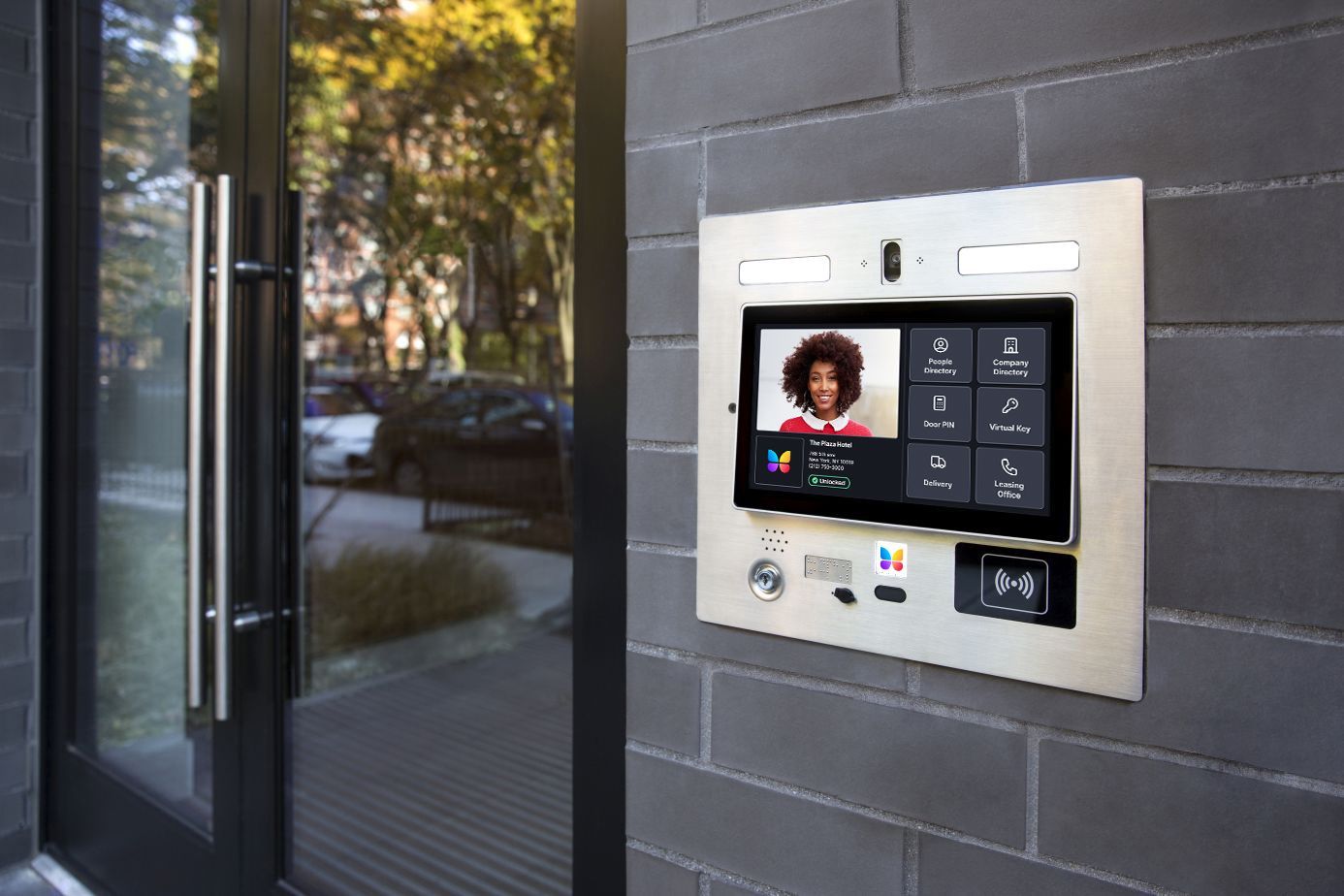 A metal smart intercom system with a touchscreen display mounted on a gray brick wall next to a glass door.