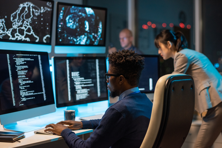 Two professionals work in a dark office at night, focused on computer screens displaying code and digital maps.