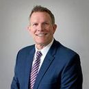 A professional headshot of a man smiling, wearing a navy blue suit, white shirt, and a purple patterned tie.