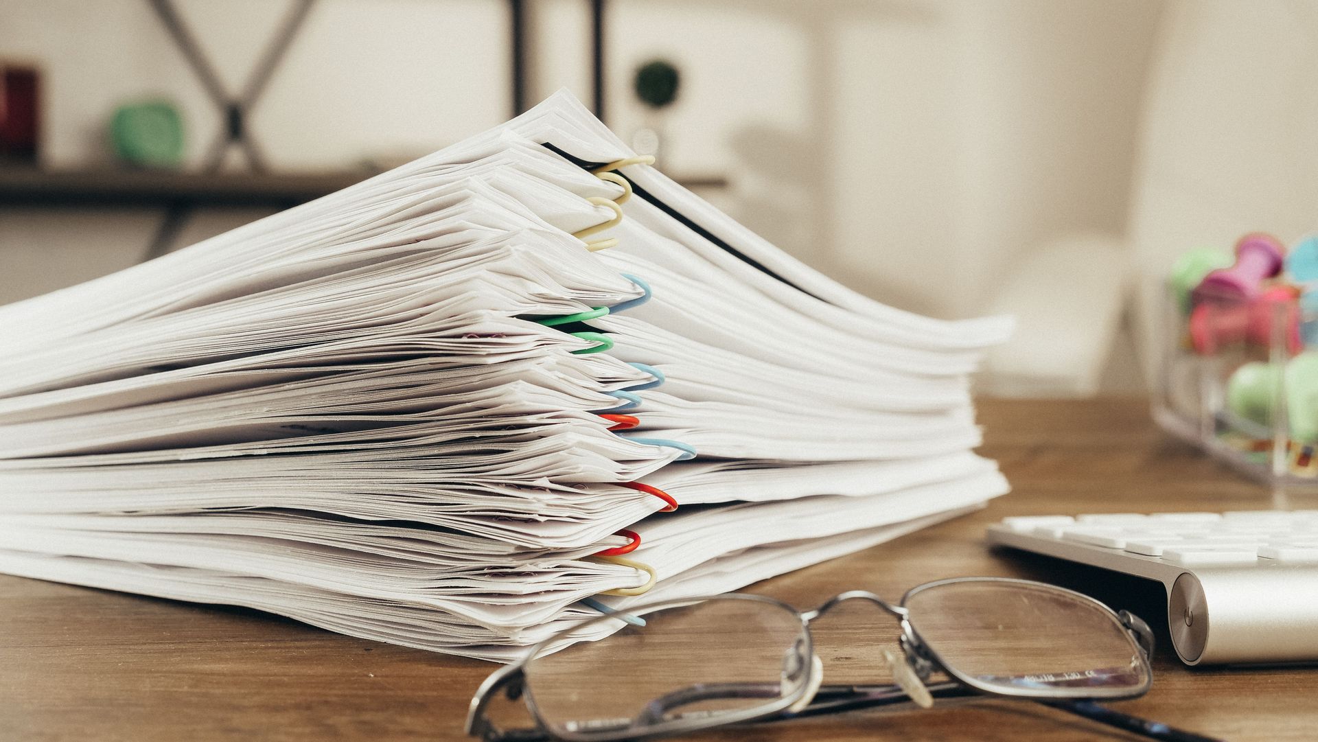 A tall stack of clipped policy and compliance paperwork beside reading glasses on an office desk.