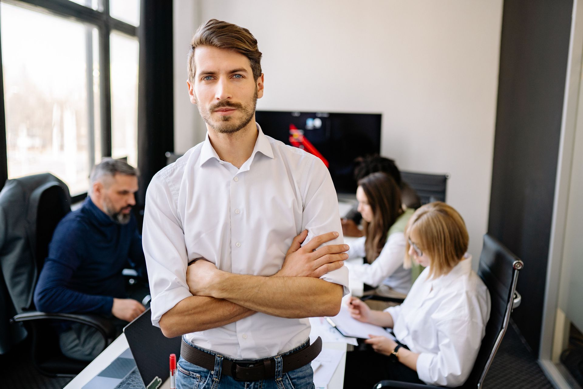 A confident office manager standing in a bright modern workplace with colleagues working in the background.