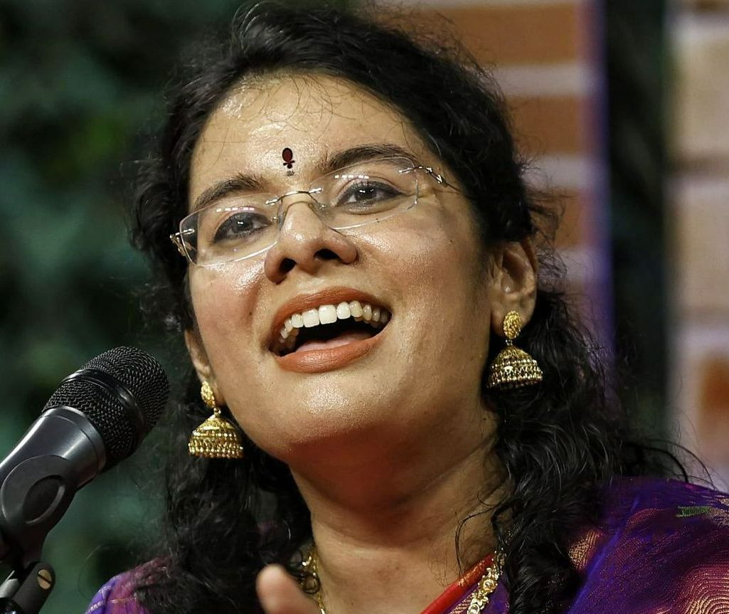 Woman singing into a microphone, wearing glasses and gold earrings, with a purple and red sari.