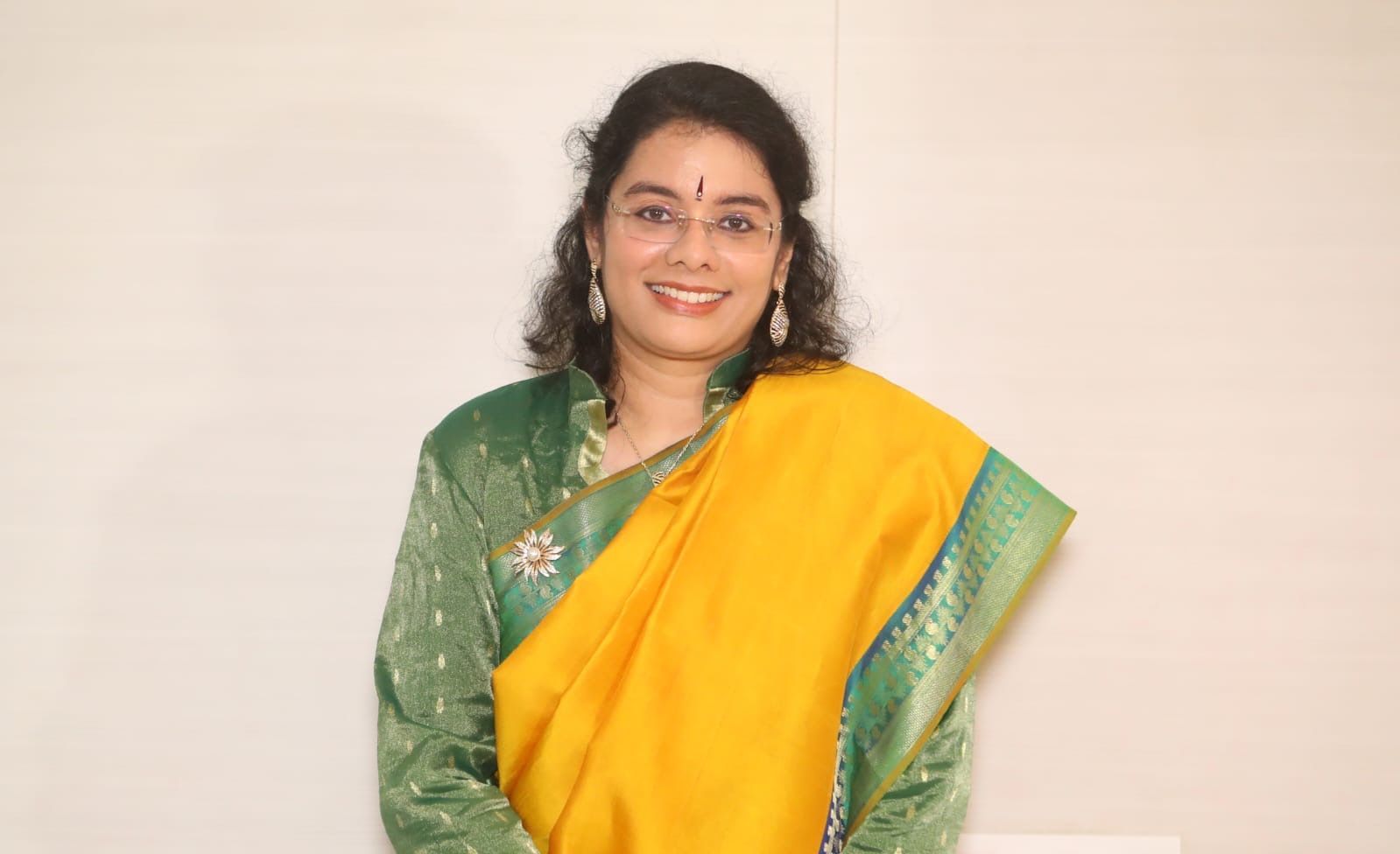 Woman in green and yellow sari smiles at the camera, posing in front of a white wall.
