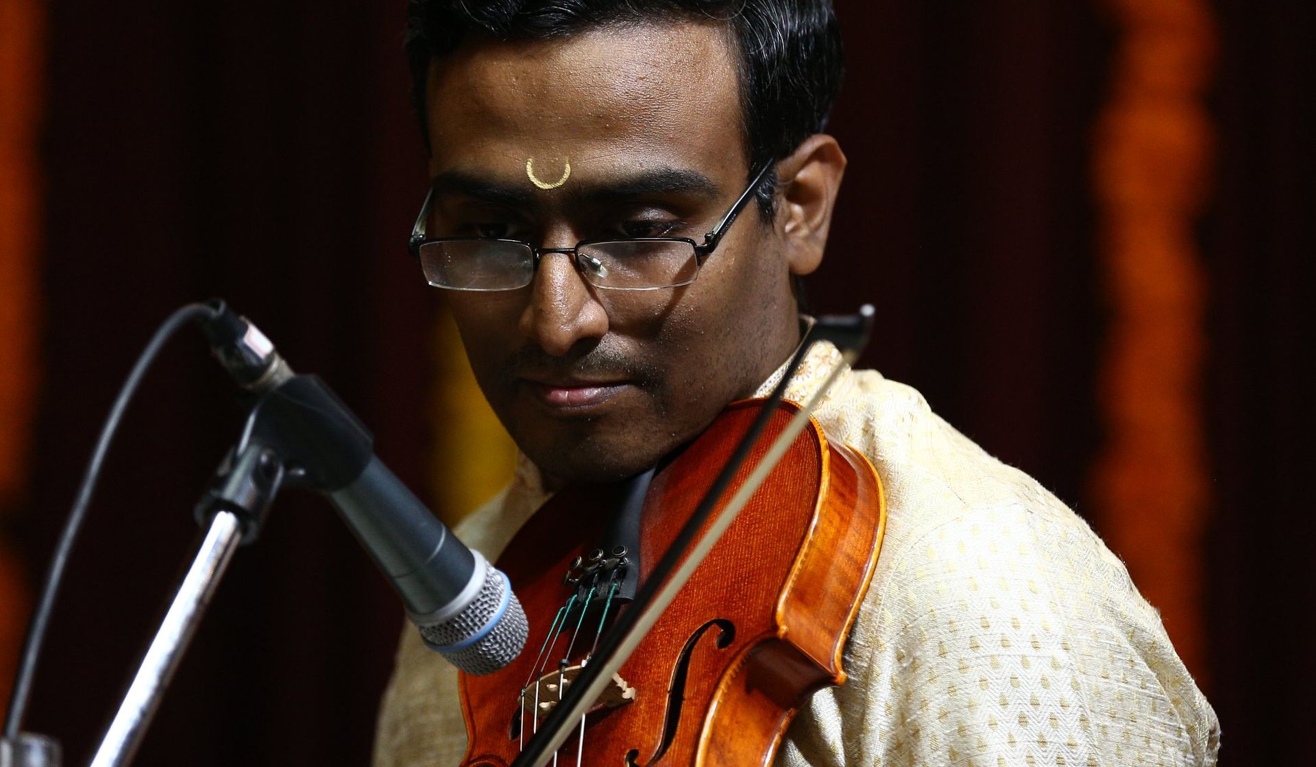 Man playing a violin, looking down, with a microphone. He wears glasses and a cream-colored shirt.