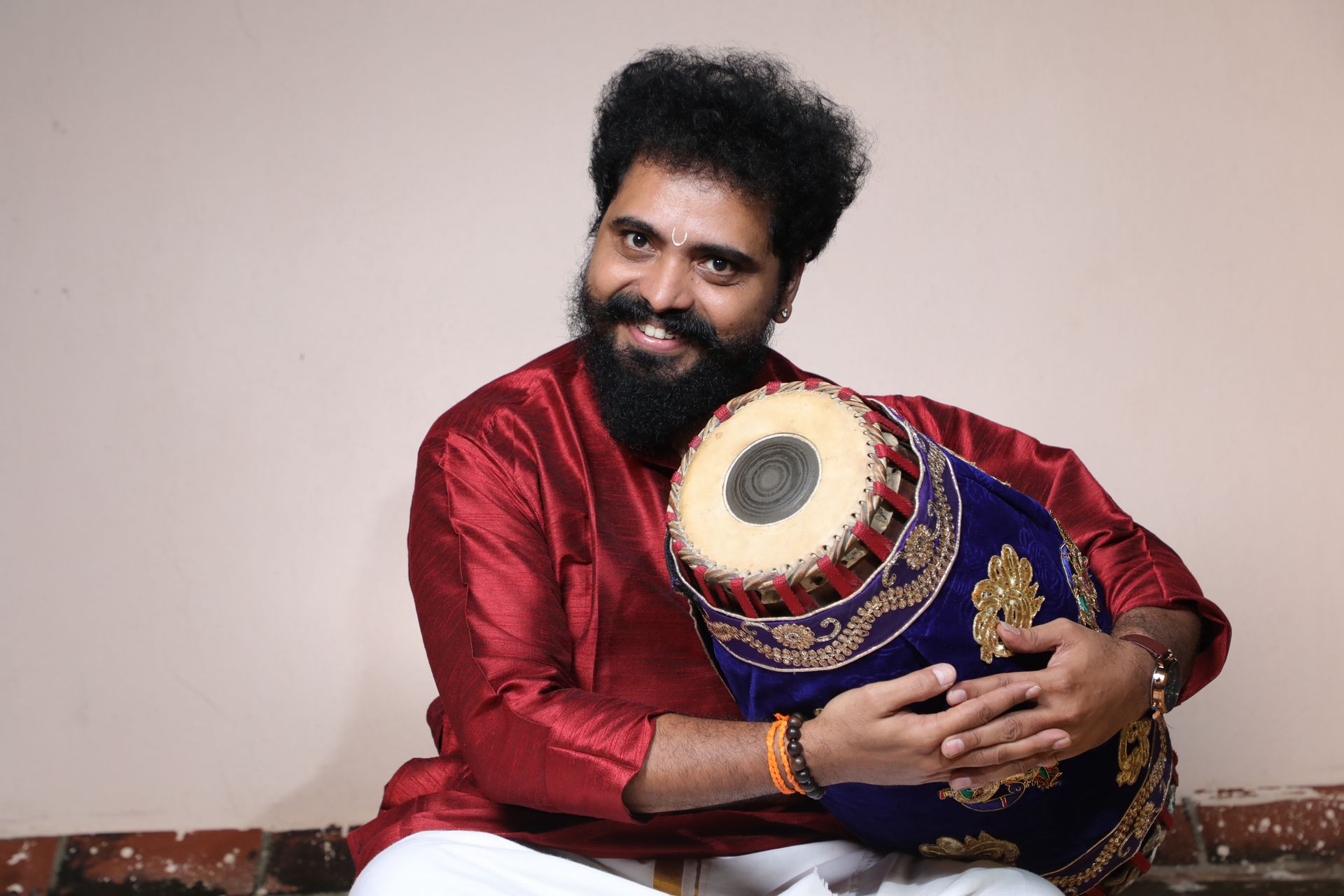 A smiling man with a beard, in red, holds a mridangam, seated in front of a beige wall.