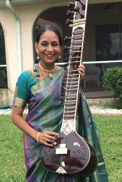 Woman in green saree smiling, holding a sitar outdoors.