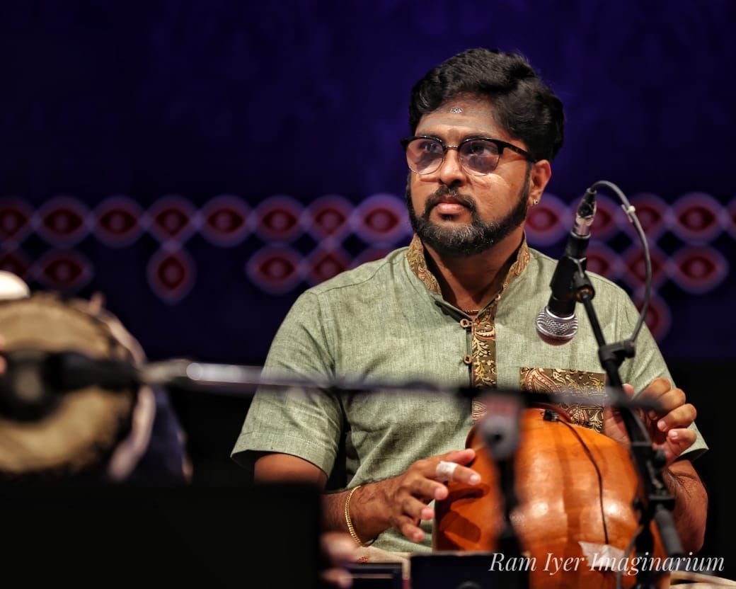Man playing a mridangam on stage, wearing glasses and a light green shirt.