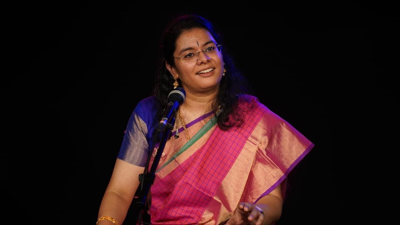 Woman in a pink and gold sari singing into a microphone, smiling on a stage.