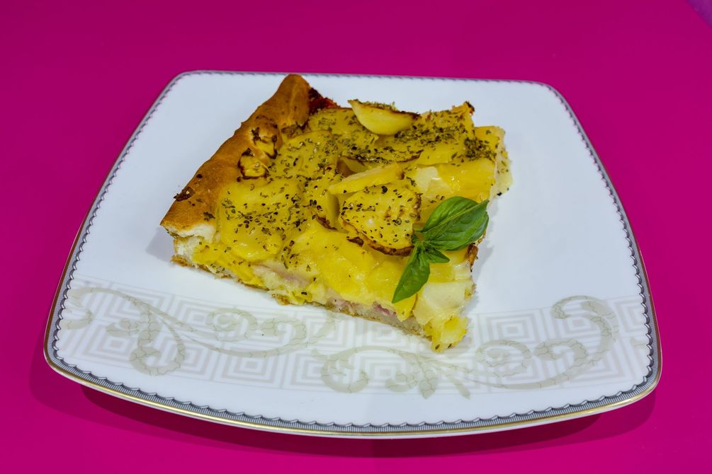 Fetta di pasta sfoglia salata gialla su un piatto bianco decorato, guarnita con erbe aromatiche, su sfondo rosa.