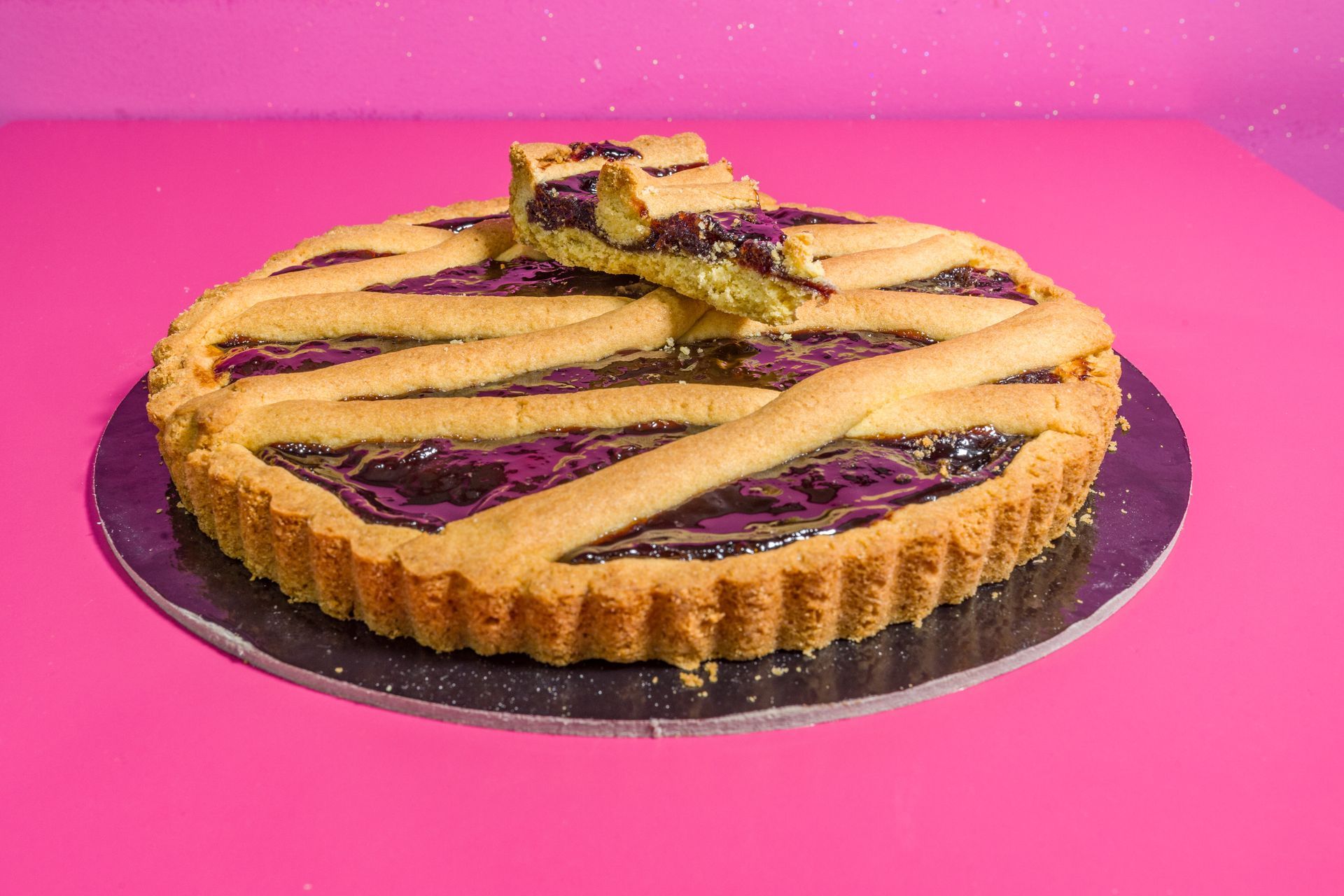 Crostata di frutti di bosco con copertura a griglia su sfondo rosa, con una fetta rimossa.