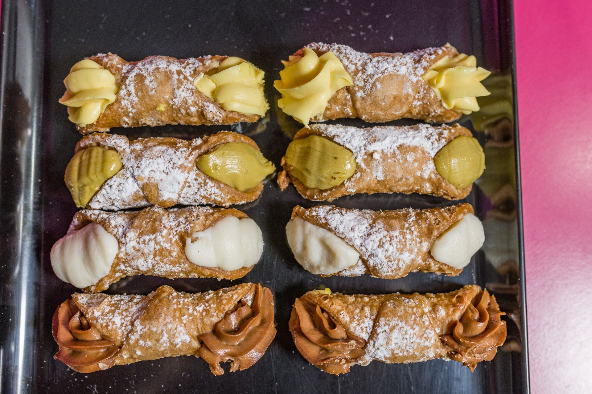 Un vassoio di otto cannoli spolverati di zucchero a velo e farciti con crema, nei gusti giallo, bianco e marrone.