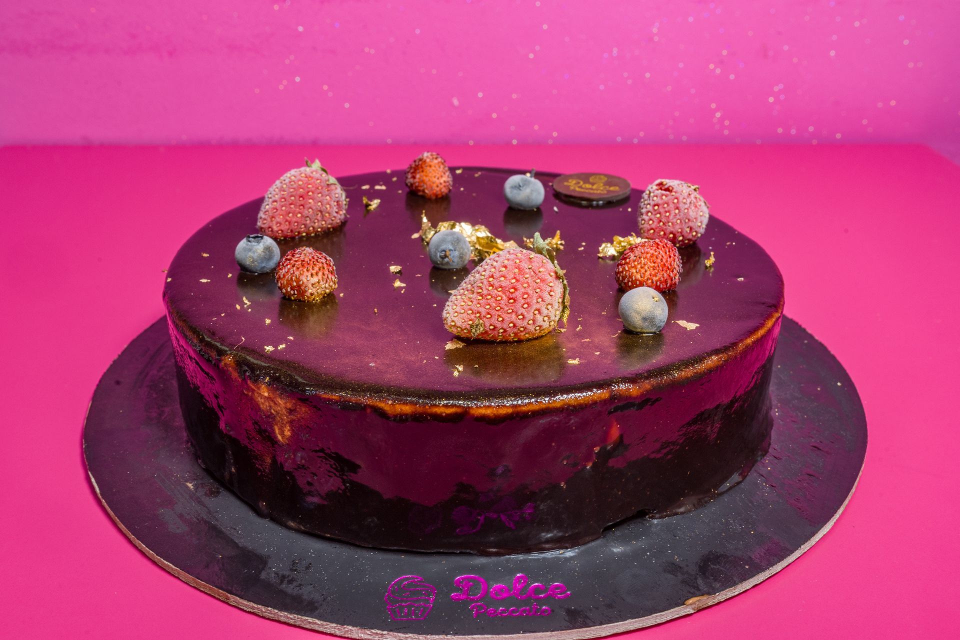 Torta al cioccolato con glassa viola, decorata con fragole e frutti di bosco su sfondo rosa.