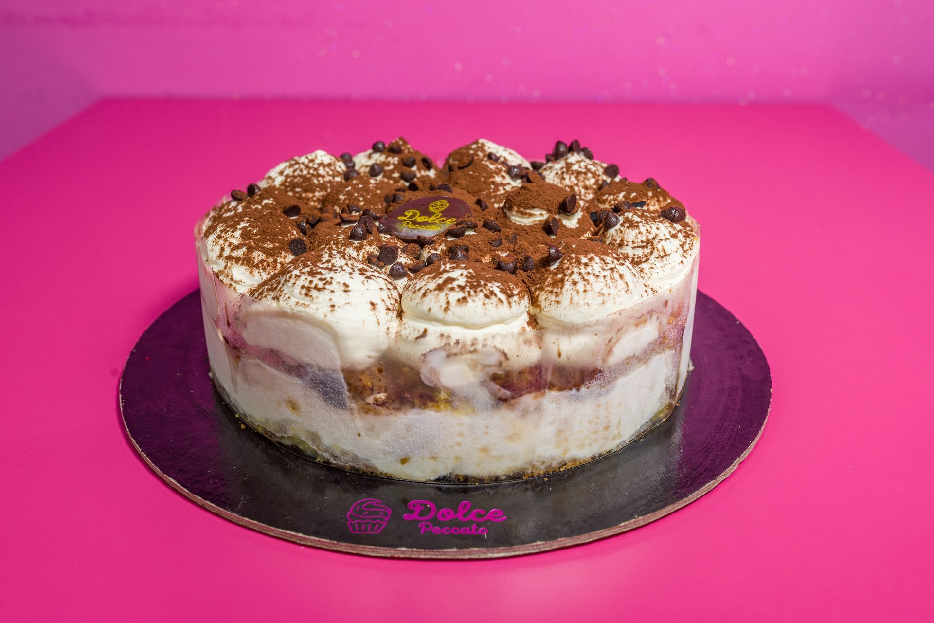 Torta rotonda a strati con glassa bianca e scaglie di cioccolato su un vassoio nero su sfondo rosa acceso.