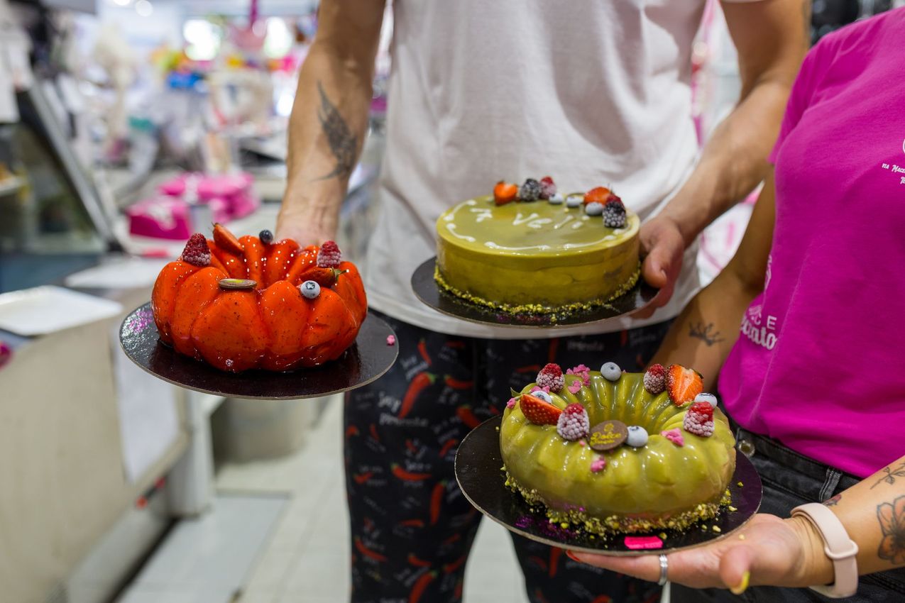 Una persona tiene in mano tre torte colorate e decorate su vassoi neri in un ambiente di negozio.