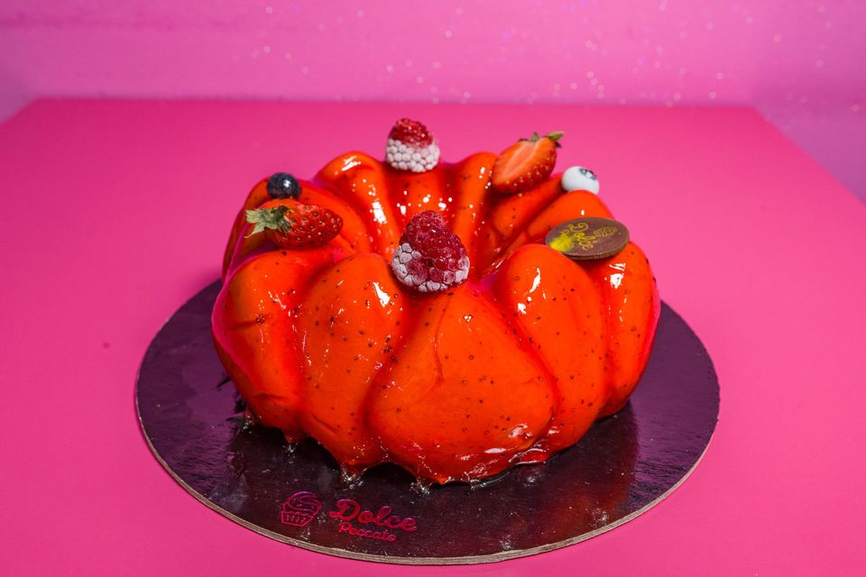Torta arancione a forma di zucca con fragole, servita su un piatto nero su sfondo rosa.