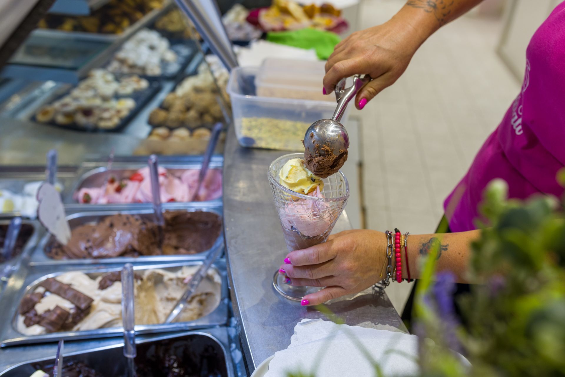 Gelato servito a mano in una coppetta al banco dei dessert, tra vassoi di dolci surgelati.