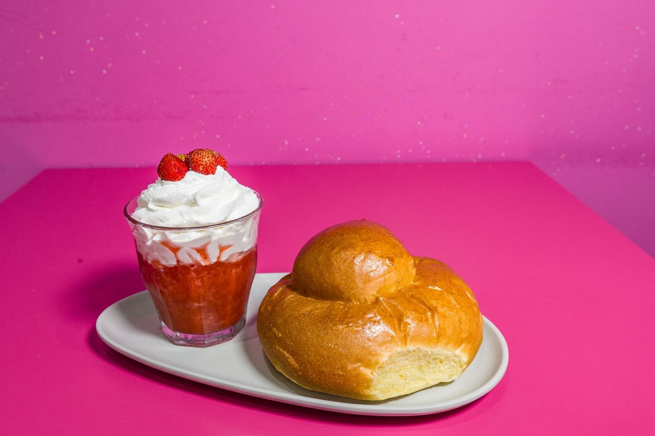 Parfait di dessert con fragole e panna montata accanto a un panino glassato su un piatto bianco su sfondo rosa.