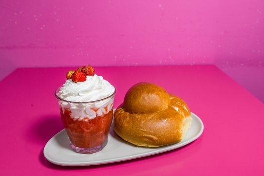 Dessert alla fragola in un bicchiere con panna montata e ciliegie, accompagnato da un panino glassato su un piatto bianco.