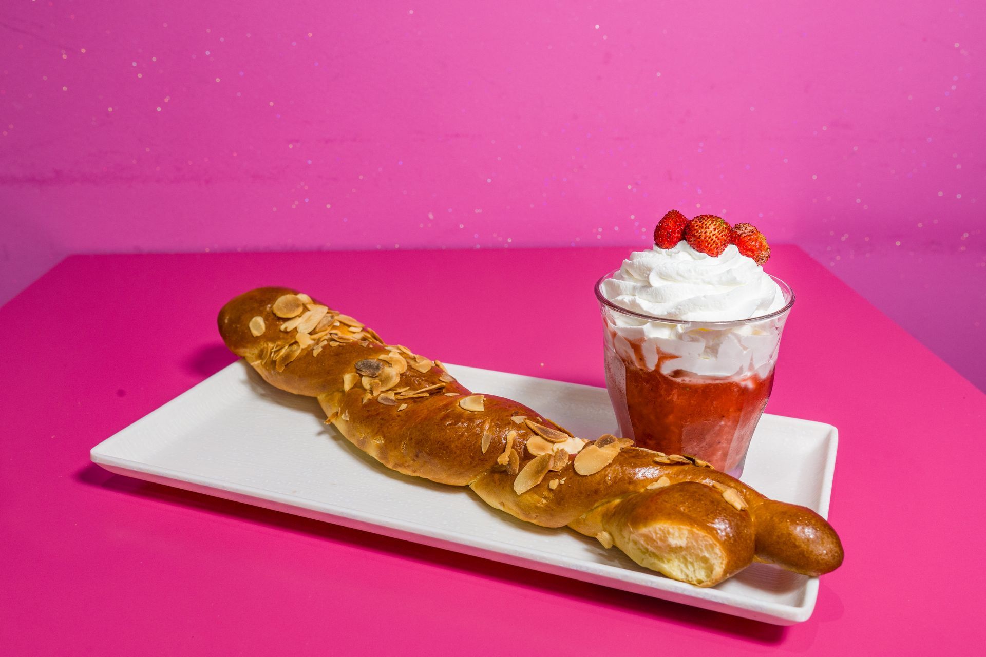 Vassoio bianco con una lunga sfoglia glassata e un dessert alla fragola guarnito con panna montata su sfondo rosa.