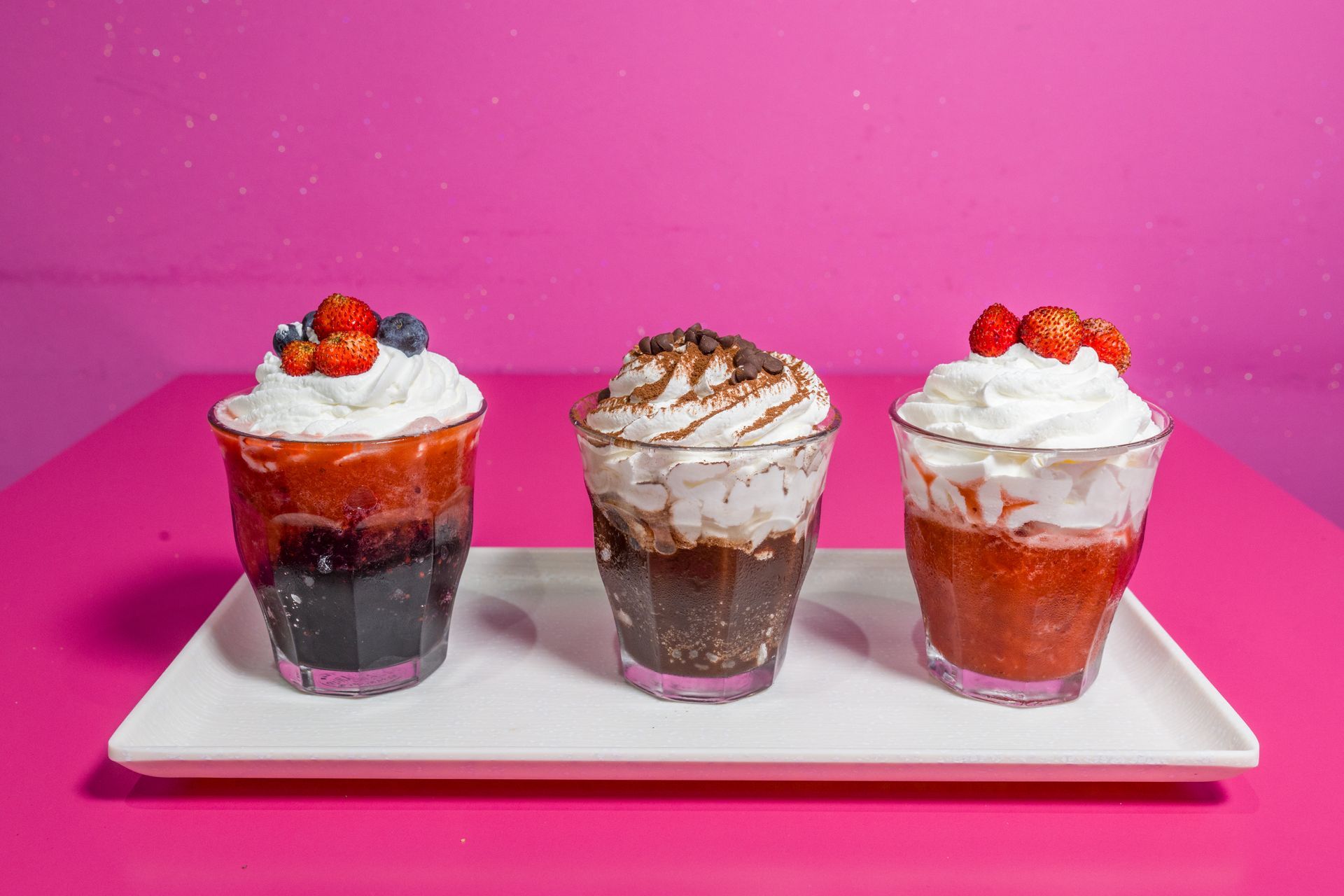 Tre parfait di dessert con panna montata e frutti di bosco su un vassoio bianco su sfondo rosa.