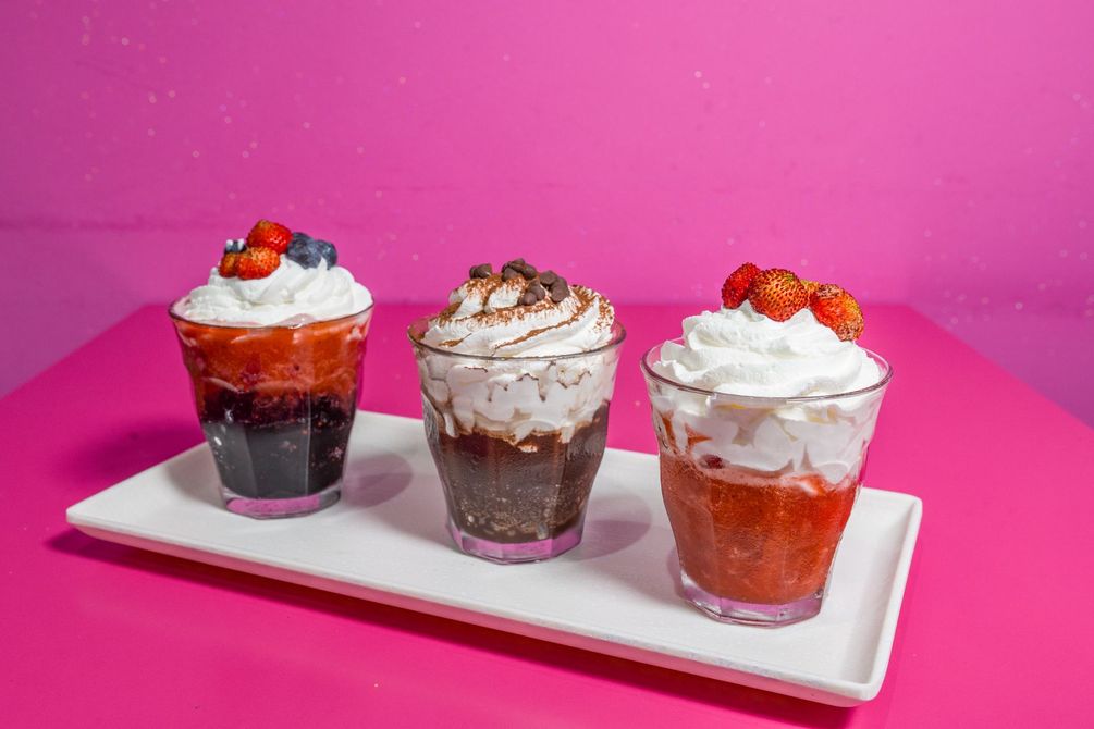 Tre coppette da dessert a strati, guarnite con panna montata e frutti di bosco, disposte su un vassoio bianco su sfondo rosa.