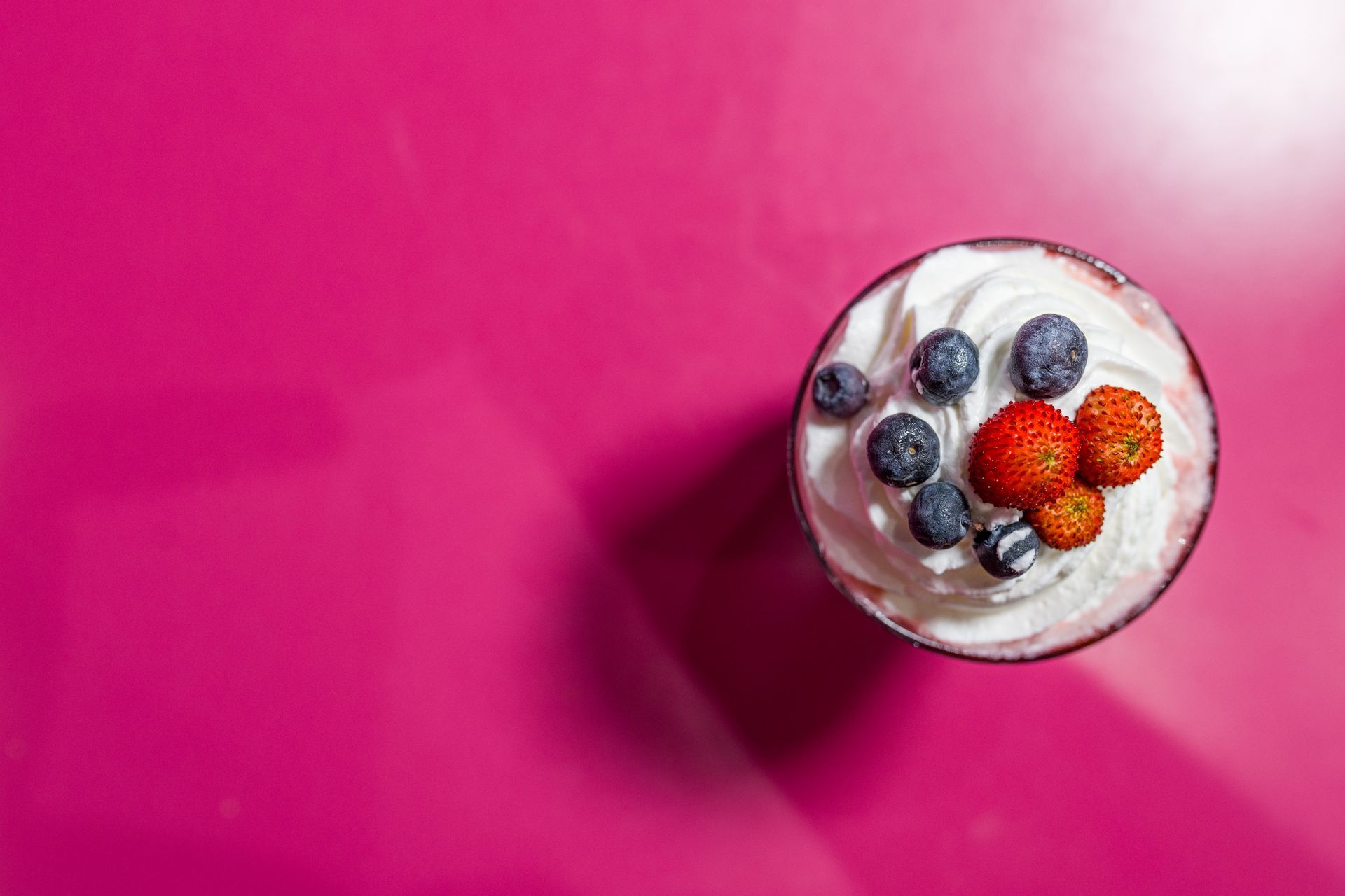 Bicchiere di yogurt guarnito con mirtilli e lamponi su uno sfondo rosa acceso.