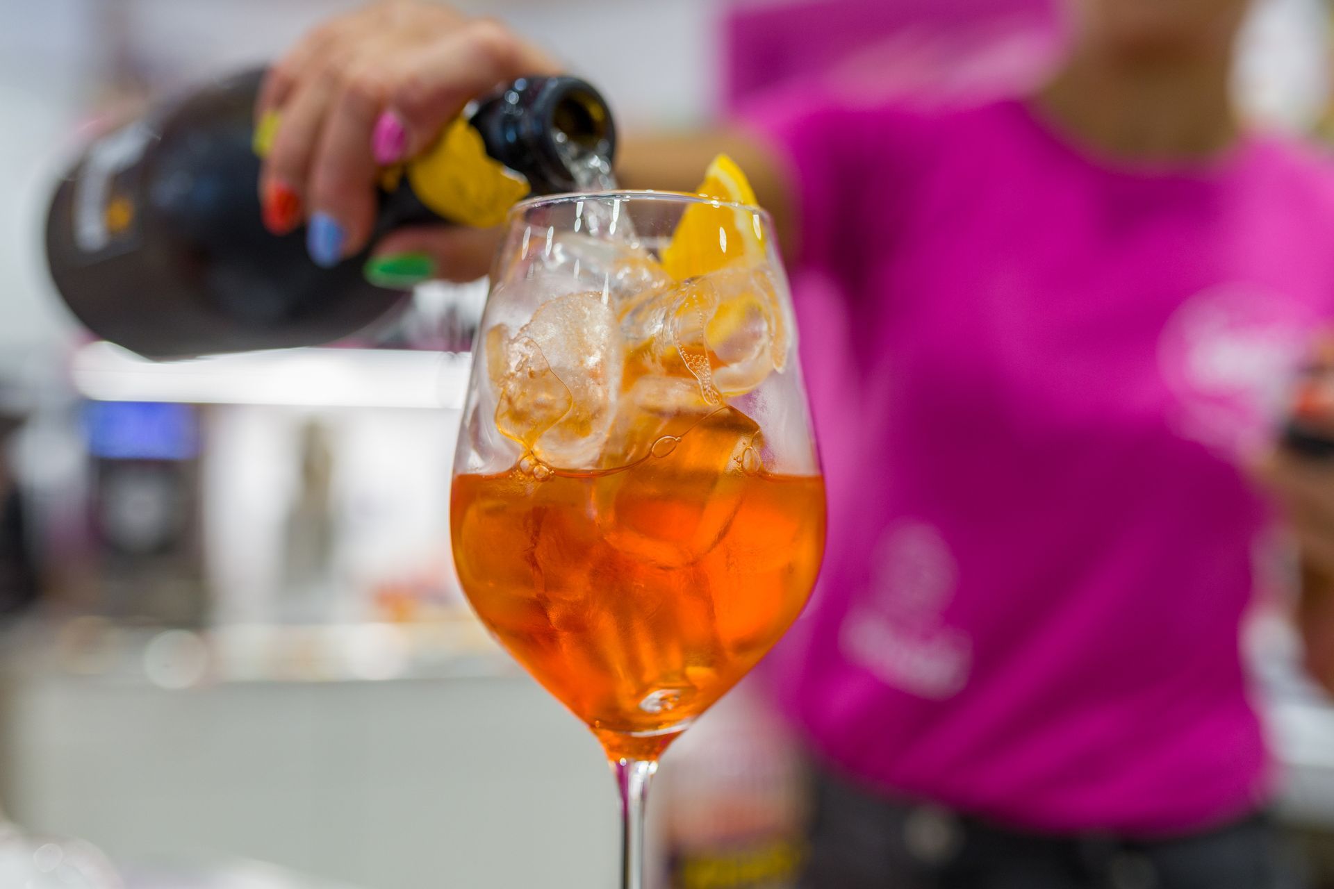 Una mano versa una bevanda all'arancia sul ghiaccio in un calice da vino, con una persona sfocata con una camicia rosa sullo sfondo.