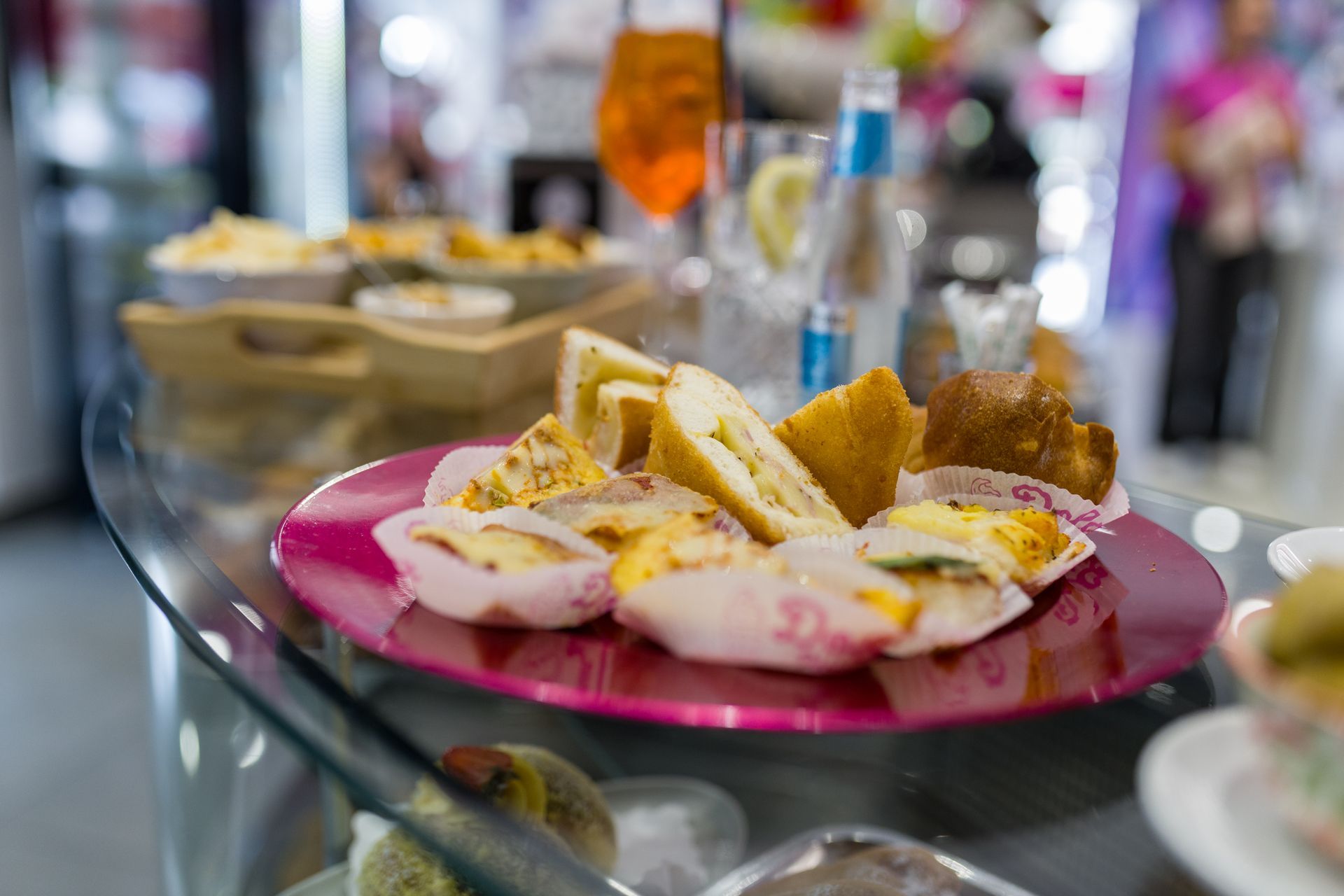 Piatto rosa con panini e pasticcini su un tavolo di vetro in un'ambientazione da caffè sfocata.