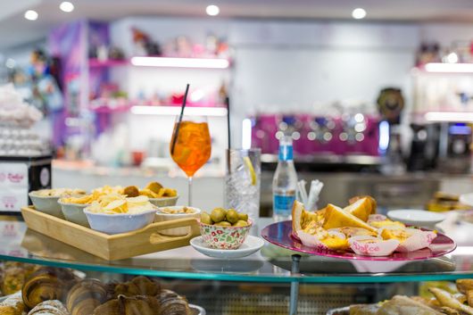 Bancone buffet con assortimento di antipasti, pasticcini e una bevanda all'arancia appesa, in un luminoso ambiente da caffetteria.