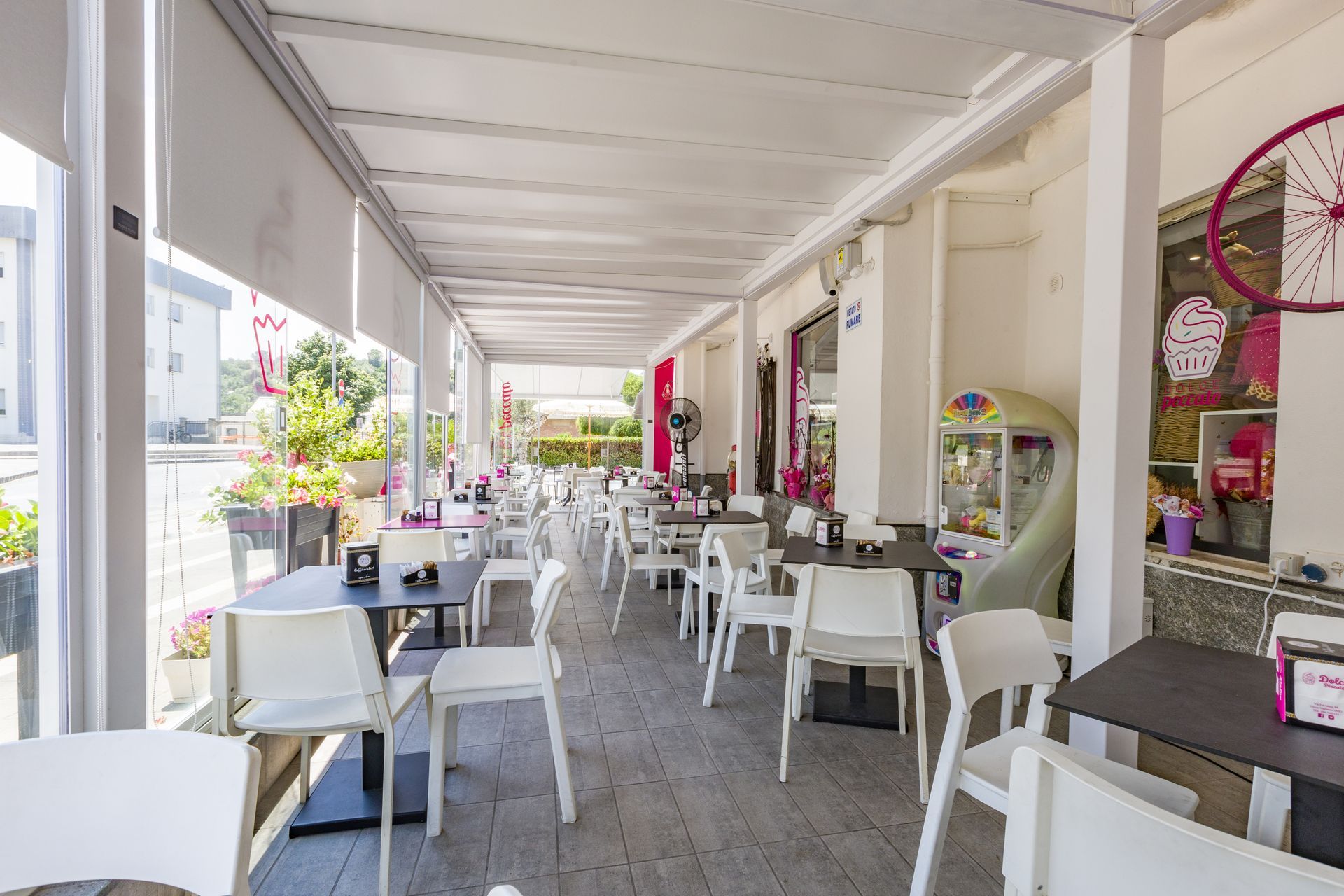 Luminoso dehor del caffè con tavolini e sedie bianchi sotto un portico coperto, decorato con fiori rosa.