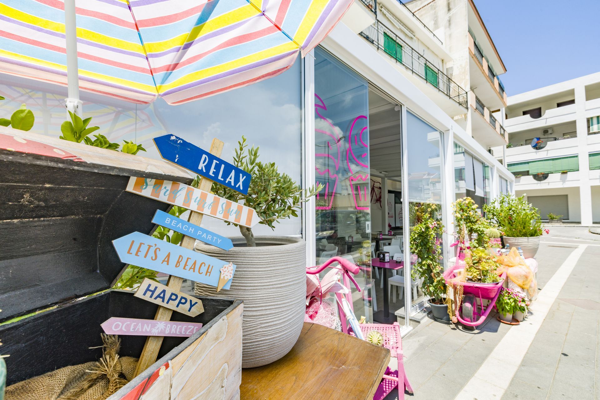 Ingresso colorato di un caffè all'aperto con ombrellone a righe, bicicletta rosa, piante in vaso e insegna con la scritta 