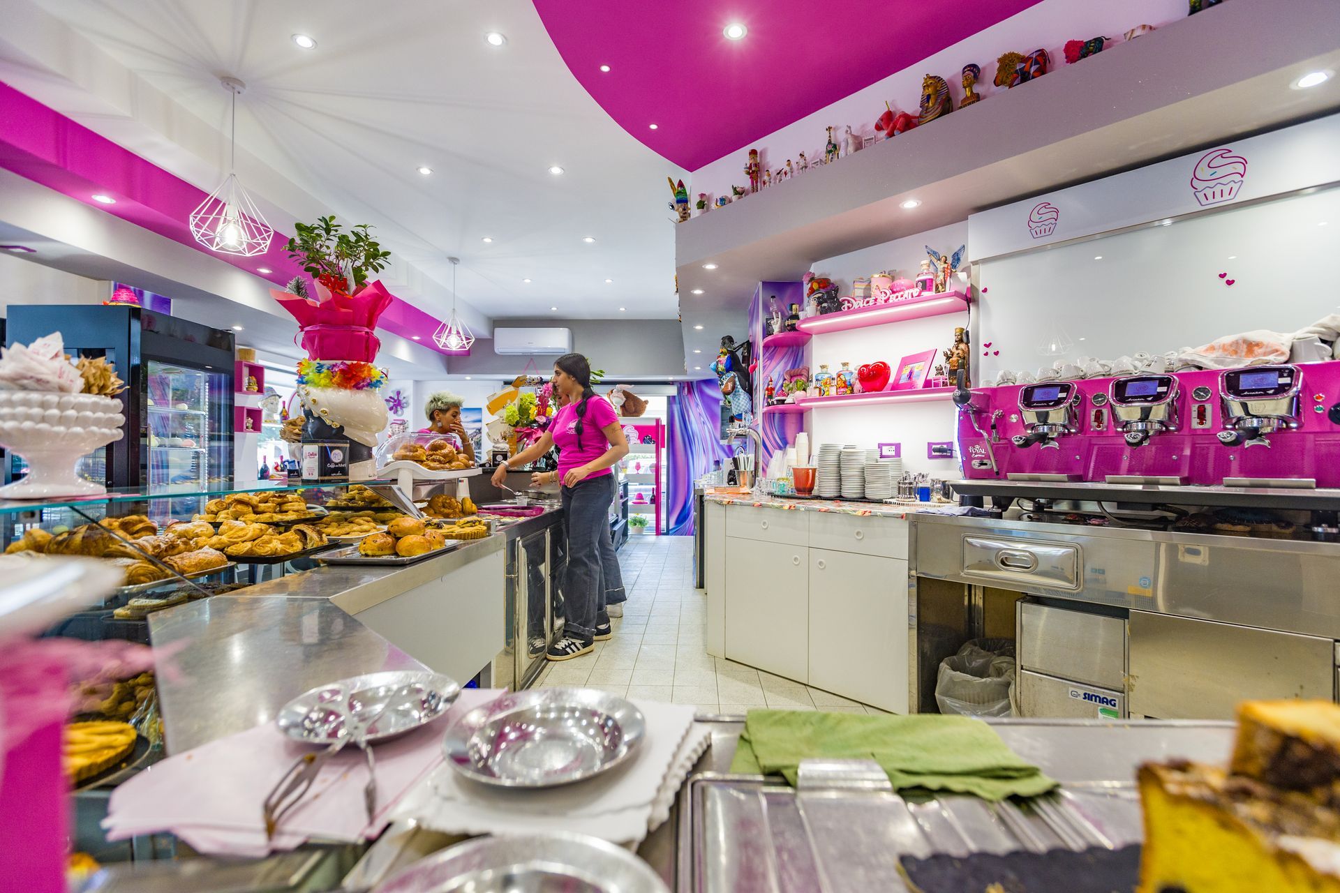 Bancone luminoso della pasticceria con pasticcini, palloncini rosa e un membro dello staff all'interno di un negozio decorato.