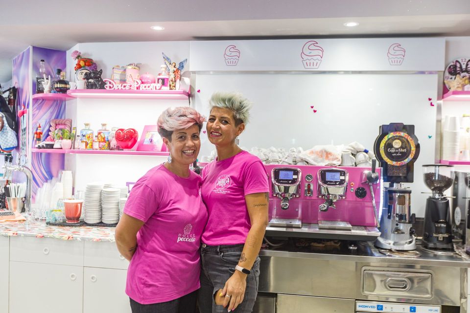 Due persone con indosso camicie rosa sono in piedi dietro il bancone di una caffetteria, dove si trovano una macchina per il caffè espresso rosa e una vetrina di dolci.