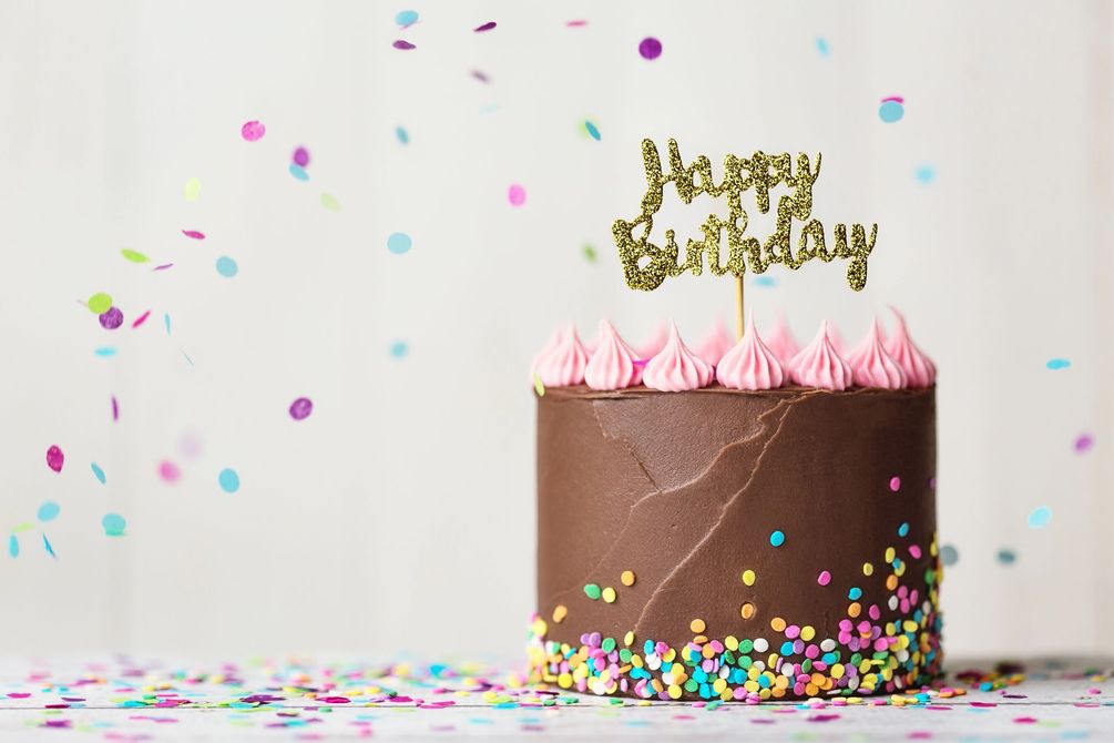 Torta di compleanno al cioccolato con glassa rosa e decorazione dorata, coriandoli sparsi su sfondo bianco.