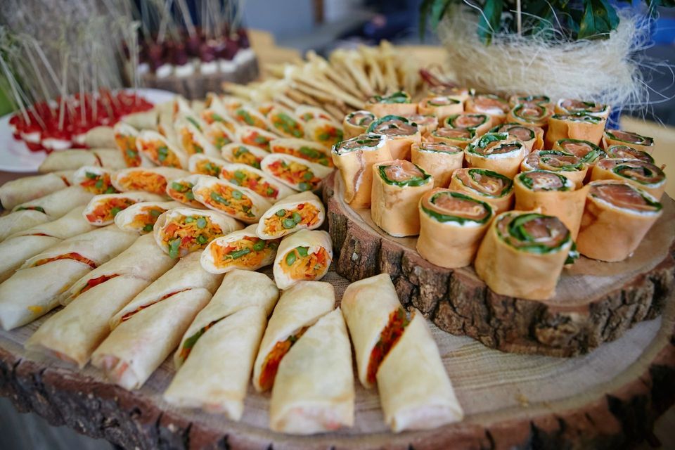 Un assortimento di stuzzichini monoporzione disposti su un piatto, tra cui piadine arrotolate e triangolini di pane alle erbe aromatiche.