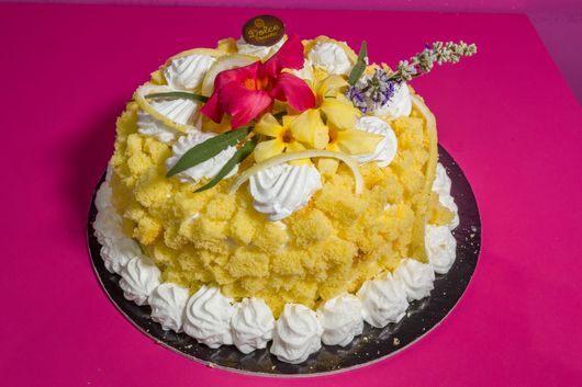 Torta gialla con decorazioni di panna montata bianca e fiori su un piatto nero su sfondo rosa.