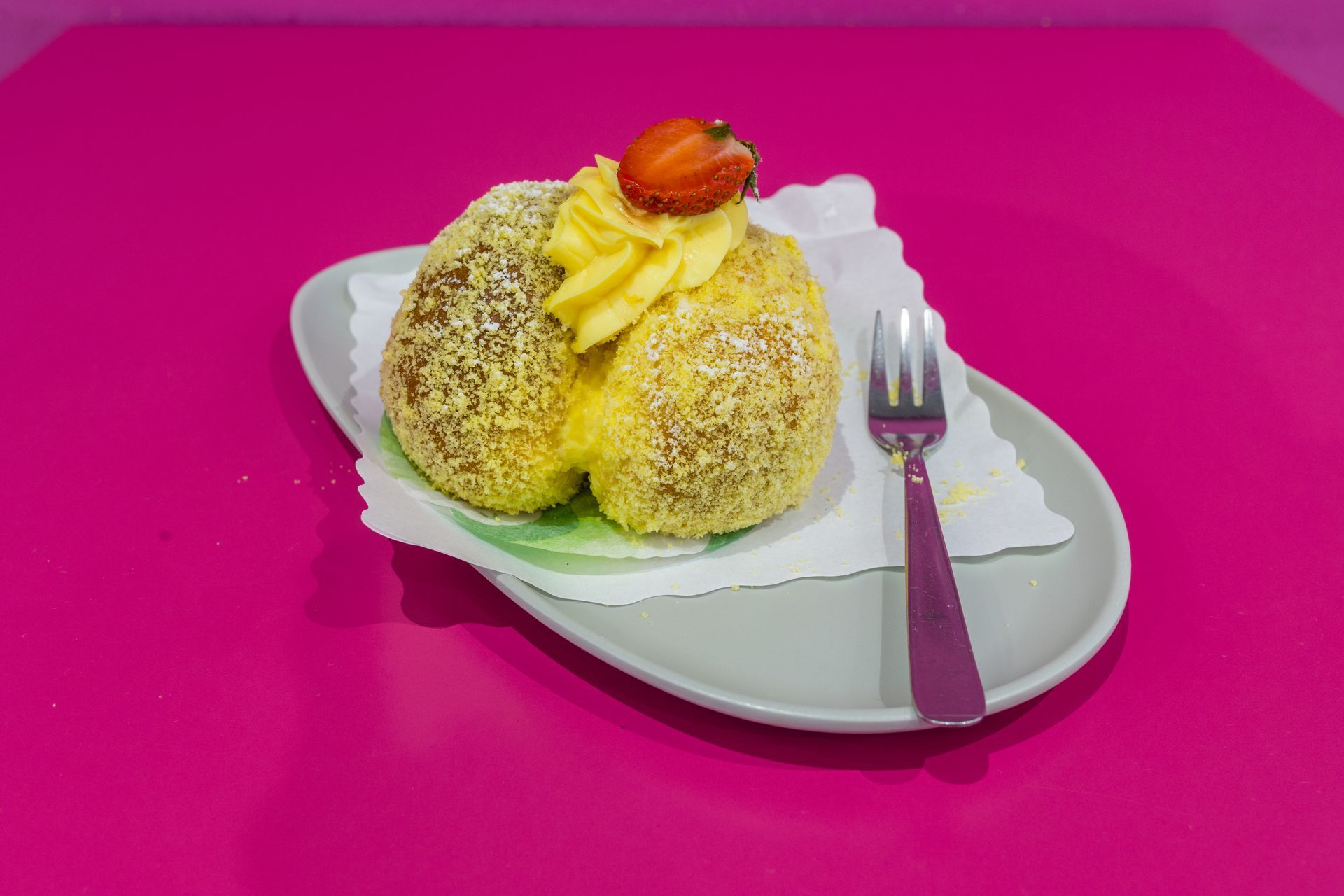 Dessert su un piatto bianco con forchetta, guarnito con panna e una fragola su sfondo rosa.