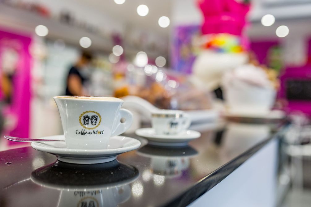 Una tazza di caffè su un piattino al bancone lucido di un bar, con altre tazze e luci colorate e sfocate sullo sfondo.