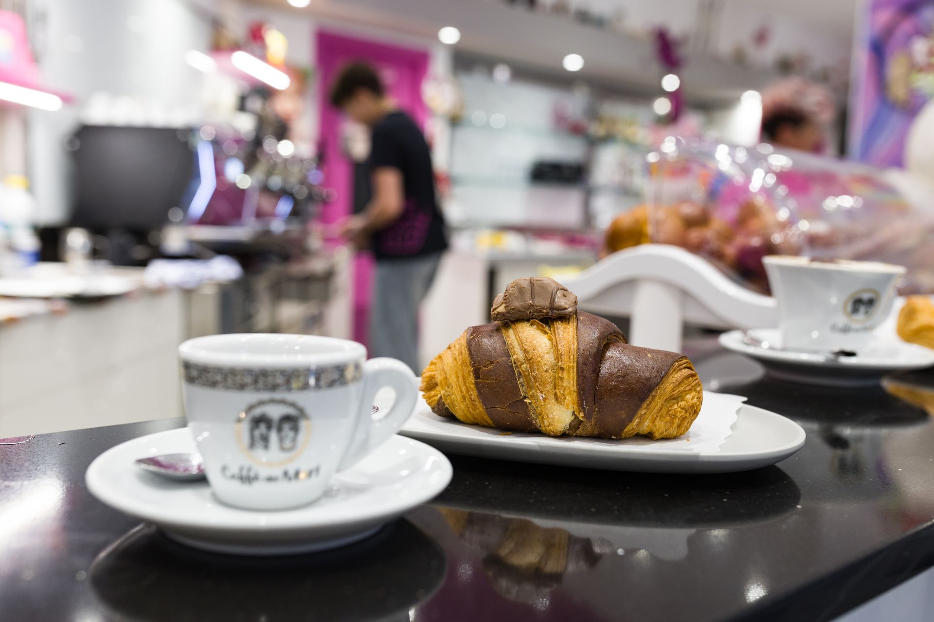 Tazze di caffè e un croissant su un tavolino di un bar, con il bancone di una panetteria sfocato sullo sfondo.