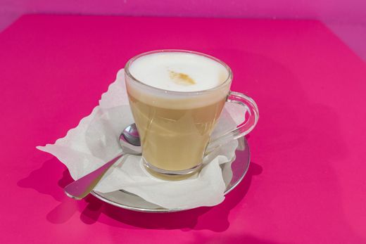 Un bicchiere di cappuccino su un piattino con cucchiaino, su una tovaglia rosa acceso.