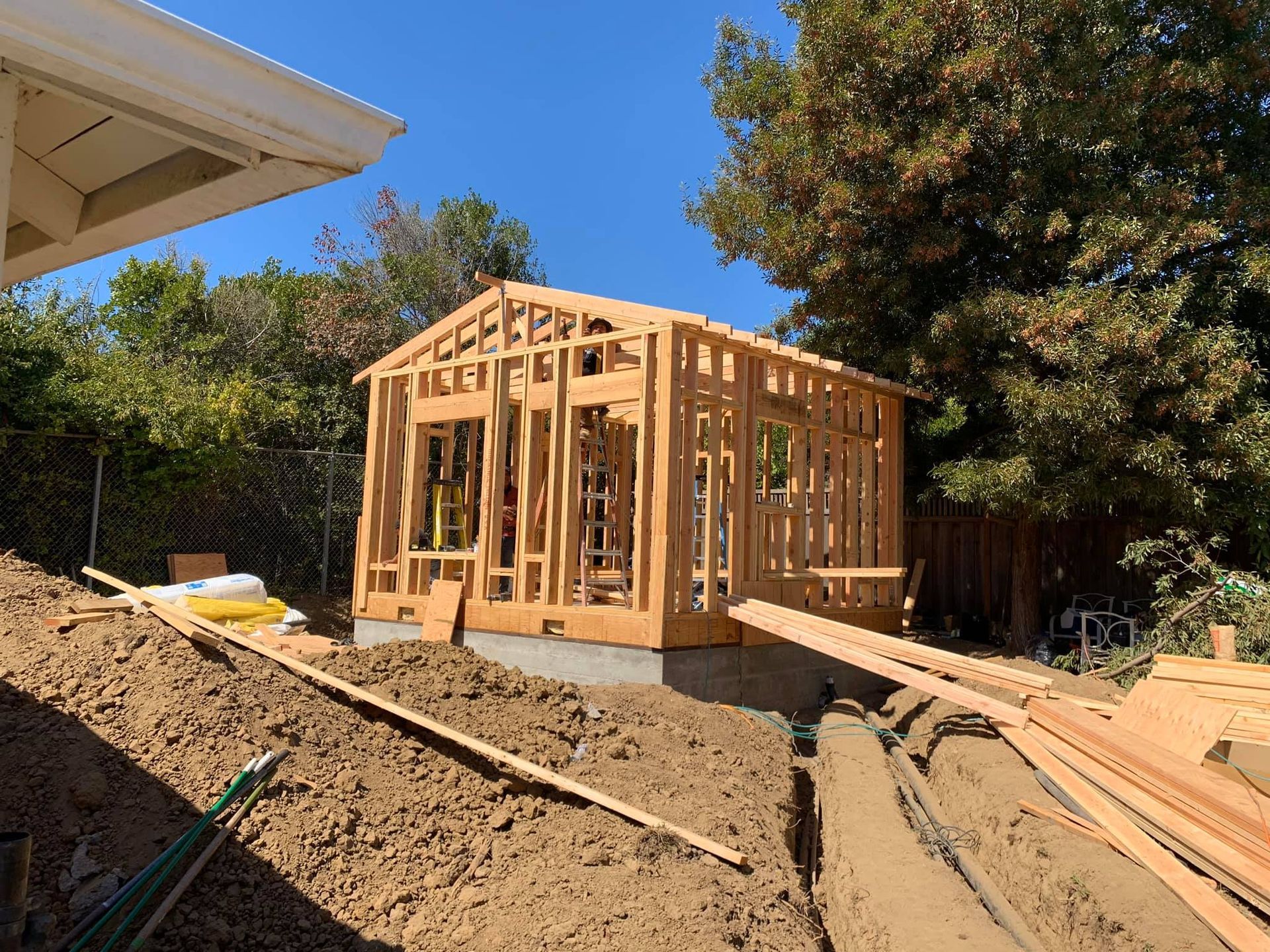 Partially built wooden house frame on a dirt construction site, with lumber stacked nearby.