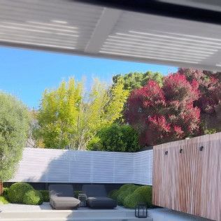Modern backyard patio with lounge chairs, white fence, and lush green and pink trees under a shaded roof