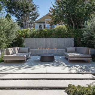 Modern patio with two gray lounge sofas, a fire pit, and a lush backyard garden view
