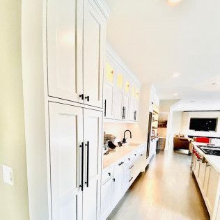 Bright modern white kitchen with tall cabinets, long counters, and a light wood floor leading to a living area