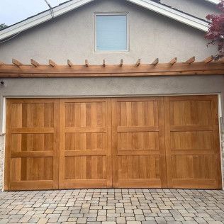 Wooden paneled garage doors on a beige house with a stone-paver driveway and pergola trim above.