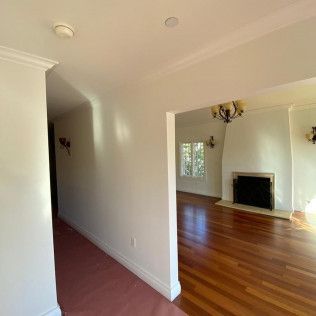Empty living room with hardwood floors, white walls, and a fireplace in a bright adjoining room