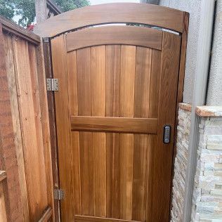 Wooden arched garden gate with a metal handle, set in a fenced outdoor walkway.