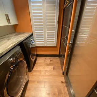 Narrow laundry room with washer, dryer, wooden floor, white shutters, and orange walls