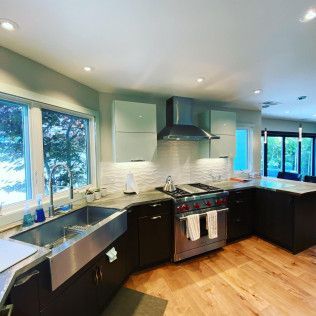 Modern kitchen with black cabinets, white backsplash, stainless stove, and hardwood floors, lit by large windows