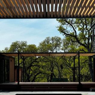 Modern patio with wooden pergola, glass railing, and trees beyond the deck